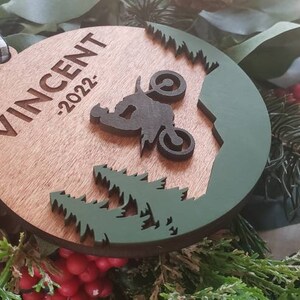 Peut inclure: Une d&eacute;coration de No&euml;l en bois avec un motif vert et marron. La d&eacute;coration repr&eacute;sente une silhouette d'une personne conduisant une moto tout-terrain dans un d&eacute;cor de for&ecirc;t. Le texte "VINCENT -2022" est grav&eacute; sur la d&eacute;coration.