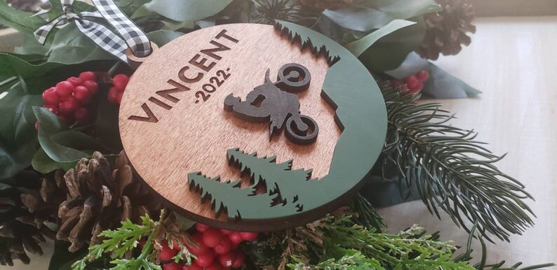 Peut inclure: Une d&eacute;coration de No&euml;l en bois avec un motif vert et marron. La d&eacute;coration repr&eacute;sente une silhouette d'une personne conduisant une moto tout-terrain dans un d&eacute;cor de for&ecirc;t. Le texte "VINCENT -2022" est grav&eacute; sur la d&eacute;coration.