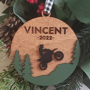 Peut inclure: Une boule de No&euml;l ronde en bois avec un motif vert et marron. La boule pr&eacute;sente une silhouette d'une personne conduisant une moto tout-terrain dans un d&eacute;cor de for&ecirc;t. La boule est personnalis&eacute;e avec le nom "Vincent" et l'ann&eacute;e "2022".