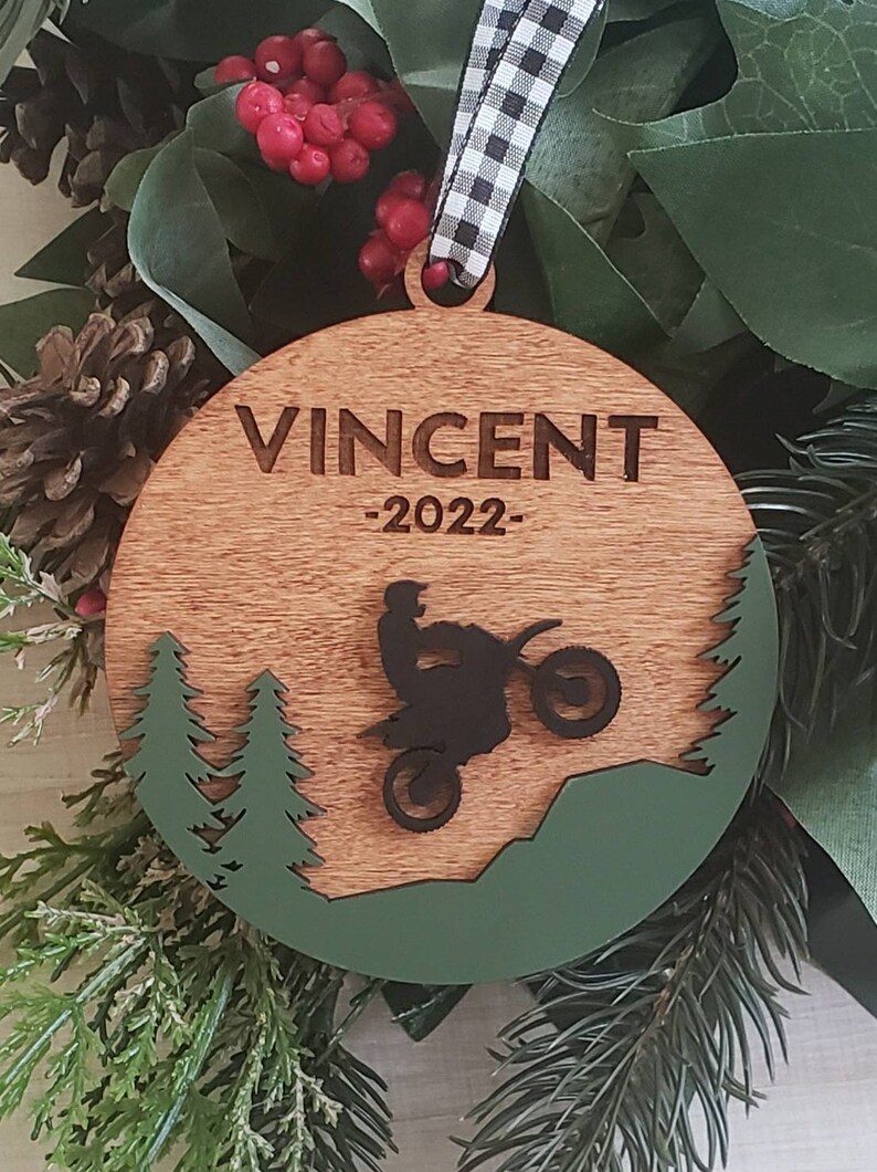 Peut inclure: Une boule de No&euml;l ronde en bois avec un motif vert et marron. La boule pr&eacute;sente une silhouette d'une personne conduisant une moto tout-terrain dans un d&eacute;cor de for&ecirc;t. La boule est personnalis&eacute;e avec le nom "Vincent" et l'ann&eacute;e "2022".