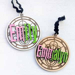 May include: Two wooden clock-themed ornaments with names and the year 2025. One ornament is white with pink and green lettering, the other is natural wood with pink and green lettering. Both have black ribbon hangers.