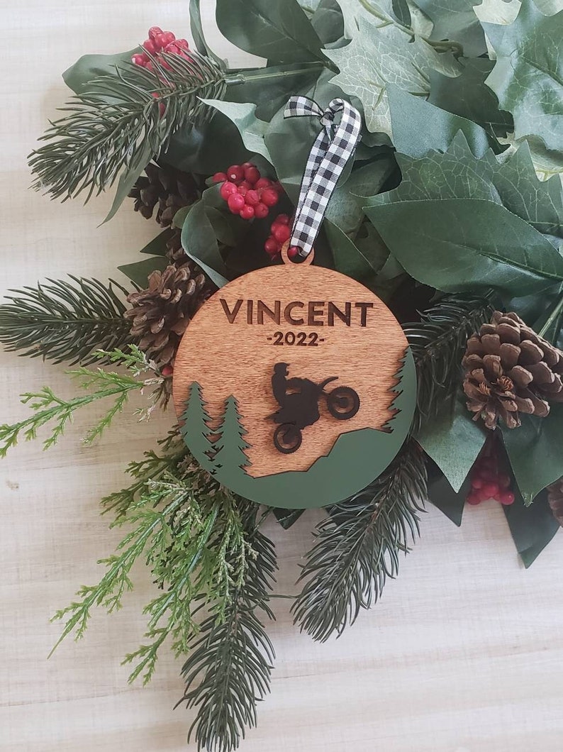 Peut inclure: Une d&eacute;coration de No&euml;l ronde en bois avec un motif vert et marron. La d&eacute;coration pr&eacute;sente une silhouette d'une personne conduisant une moto tout-terrain dans un d&eacute;cor de for&ecirc;t. Le texte "VINCENT -2022- " est grav&eacute; sur la d&eacute;coration.