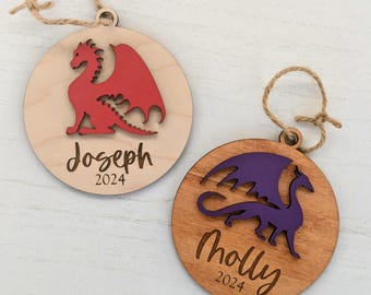 May include: Two wooden Christmas ornaments with a dragon design. The ornament on the left has a red dragon and the text "Joseph 2024". The ornament on the right has a purple dragon and the text "Molly 2024".