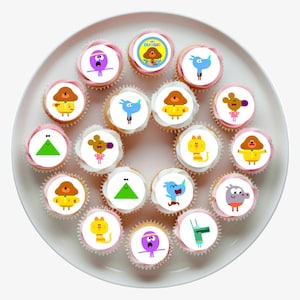 May include: A collection of cupcakes arranged on a white plate, each topped with a circular edible topper featuring colourful cartoon characters from Hey Duggee, including Duggee, Norrie, Tag, Betty, Roly, and Happy, with various expressions.