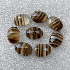 May include: Eight oval-shaped, faceted, brown and white agate gemstones. The gemstones have a striped pattern and are arranged in a circle on a white surface.