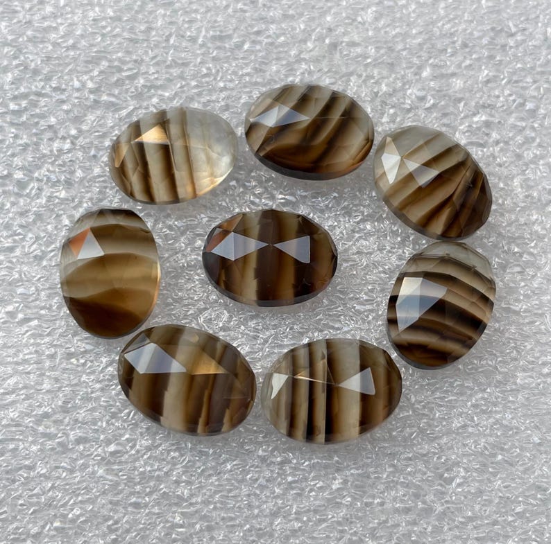 May include: Eight oval-shaped, faceted, brown and white agate gemstones. The gemstones have a striped pattern and are arranged in a circle on a white surface.