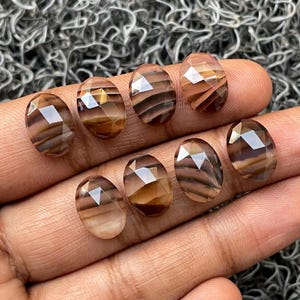 May include: Eight oval-shaped, faceted, brown and white agate gemstones. The gemstones have a striated pattern and are arranged in a row on a hand.