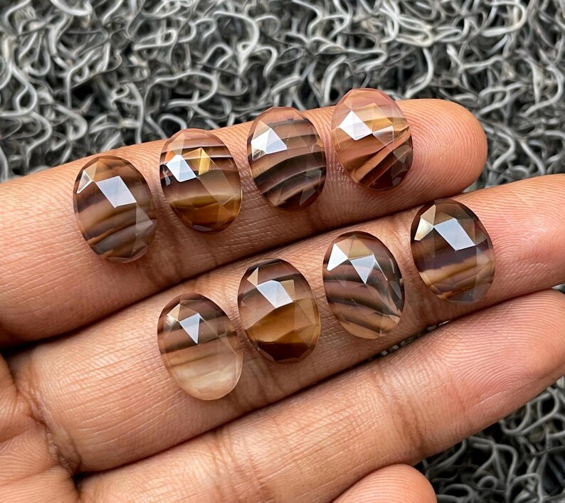 May include: Eight oval-shaped, faceted, brown and white agate gemstones. The gemstones have a striated pattern and are arranged in a row on a hand.