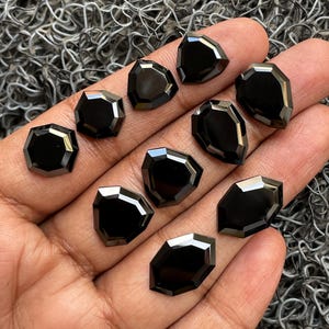 May include: A collection of black gemstone cabochons in various geometric shapes, including octagons and teardrops. The polished stones are displayed on a hand, showcasing their reflective surfaces and dark color.