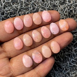 Könnte beinhalten: Eine Sammlung oval geschliffener Edelsteine in verschiedenen Rosa- und Pfirsichtönen. Die facettierten Steine werden auf einem neutralen Hintergrund präsentiert, um ihre polierten Oberflächen und Farbvariationen hervorzuheben. Ideal für die Schmuckherstellung.