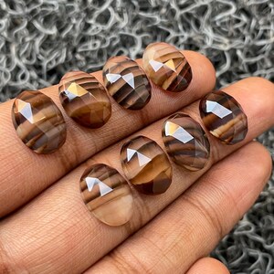 May include: Eight faceted, oval-shaped gemstones with brown and white stripes. The stones are arranged in a row on a hand.