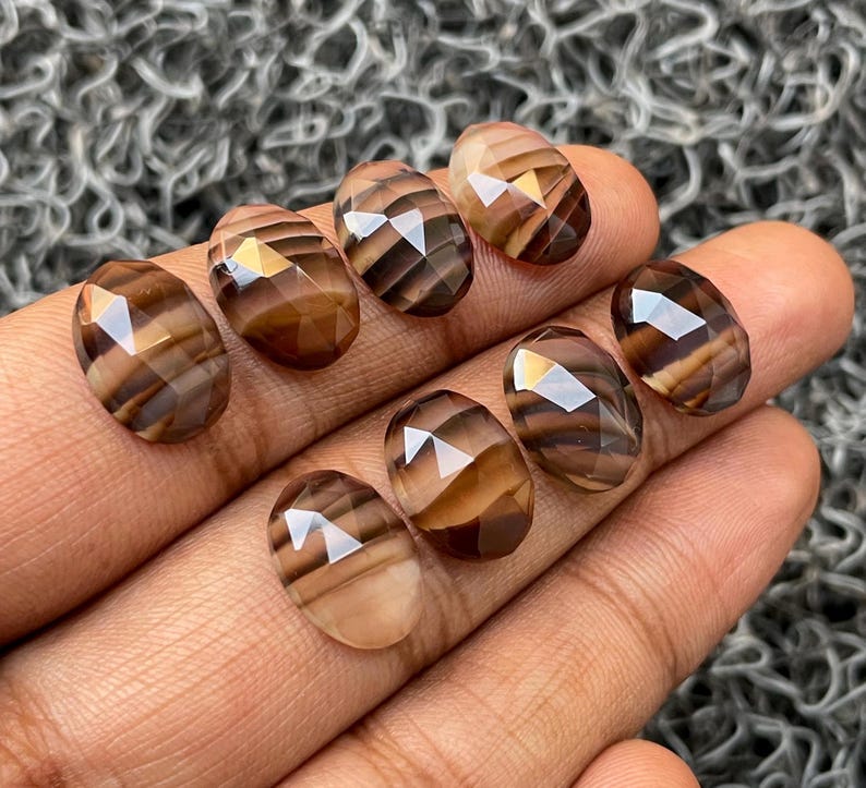 May include: Eight faceted, oval-shaped gemstones with brown and white stripes. The stones are arranged in a row on a hand.