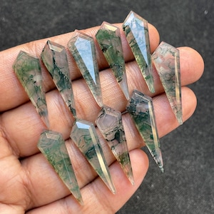 May include: A collection of ten clear, faceted gemstone pendants. Each pendant has a pointed bottom and a geometric top. The gemstones have green and pink inclusions, creating a natural, organic pattern. The pendants are held in a hand.
