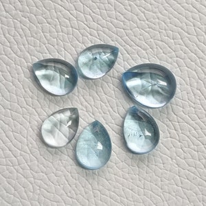 May include: Six teardrop-shaped aquamarine gemstones, ranging in size and shade of blue, on a white textured surface.