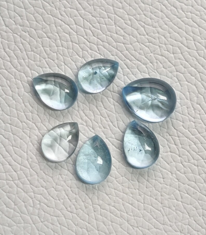 May include: Six teardrop-shaped aquamarine gemstones, ranging in size and shade of blue, on a white textured surface.