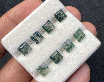 May include: Eight square-cut moss agate gemstones displayed on a white surface. The gemstones have a translucent appearance with green, moss-like inclusions. The gems are held in a hand, showcasing their size and clarity. Suitable for jewellery making.