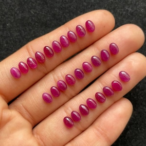 以下が含まれることがあります： 20個の楕円形のファセットカットされたルビーレッドの宝石のコレクション。宝石は手のひらに並べて置かれています。