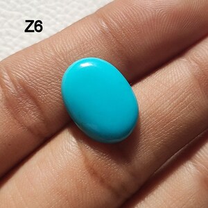 May include: A bright turquoise oval cabochon gemstone, likely turquoise, held in a hand. The stone is smooth and polished.