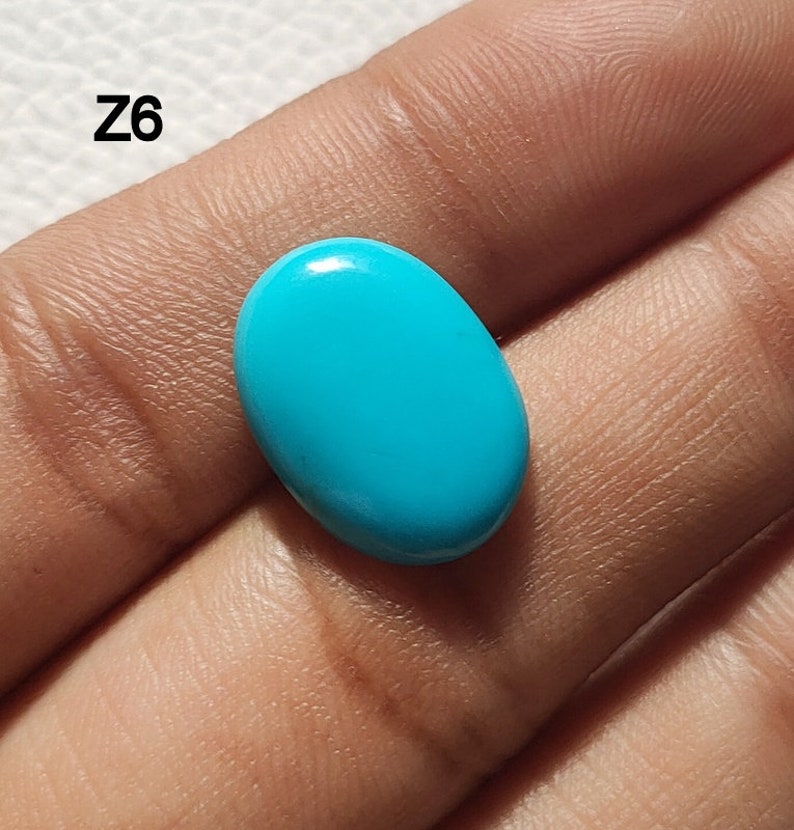 May include: A bright turquoise oval cabochon gemstone, likely turquoise, held in a hand. The stone is smooth and polished.