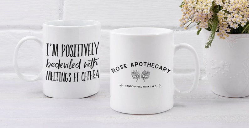 May include: Two white ceramic mugs with black text. The mug on the left says "I'm positively bedeviled with meetings et cetera". The mug on the right says "Rose Apothecary Handcrafted with care".