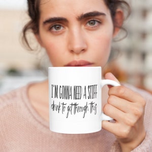 May include: White ceramic coffee mug with black text that reads "I'm gonna need a stiff drink to get through this".