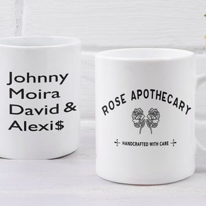 May include: Two white ceramic mugs with black text. The mug on the left says "Johnny Moira David & Alexis". The mug on the right says "Rose Apothecary" with a stylized image of two roses and the text "Handcrafted with care".
