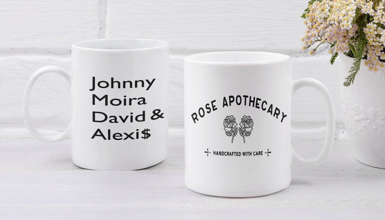 May include: Two white ceramic mugs with black text. The mug on the left says "Johnny Moira David & Alexis". The mug on the right says "Rose Apothecary" with a stylized image of two roses and the text "Handcrafted with care".