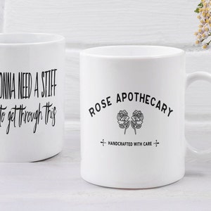May include: Two white ceramic mugs with black text and designs. The mug on the left says "I'm gonna need a stiff drink to get through this". The mug on the right says "Rose Apothecary" with a hand-drawn rose design and the text "Handcrafted with care".