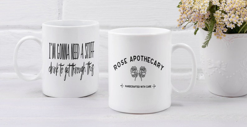 May include: Two white ceramic mugs with black text and designs. The mug on the left says "I'm gonna need a stiff drink to get through this". The mug on the right says "Rose Apothecary" with a hand-drawn rose design and the text "Handcrafted with care".