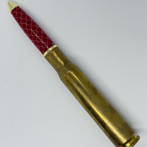 Può includere: Una penna a sfera realizzata con un bossolo di proiettile in ottone. Il fusto superiore presenta una finitura rosso vivace con un motivo a nido d'ape bianco, accentuato da una punta dorata e un sottile anello dorato che separa la sezione rossa dalla base.