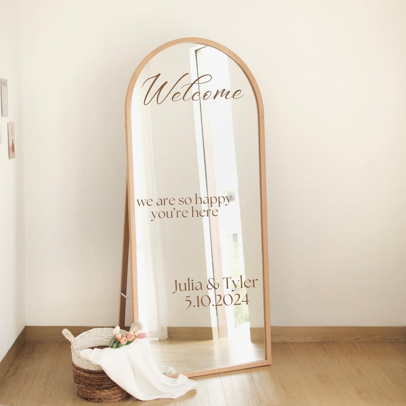 Op de afbeelding: Een houten boogspiegel met een welkomstbord en de tekst "we are so happy you're here" en "Julia & Tyler 5.10.2024".