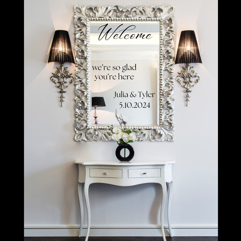 Op de afbeelding: Een zilveren, sierlijke spiegel met een welkomstbord waarop staat "Welcome we're so glad you're here Julia & Tyler 5.10.2024". De spiegel hangt aan een witte muur boven een witte tafel met twee laden.