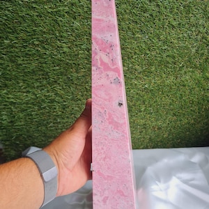 May include: A tall, pink obelisk with a pointed top. The stone features a marbled pattern of light and dark pink with black speckles. The obelisk is held in a hand, with a silver watch band visible.