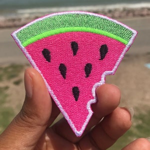May include: A pink watermelon slice with a bite taken out of it. The watermelon is embroidered with white stitching and has black seeds. The watermelon is on a green background with a white border.