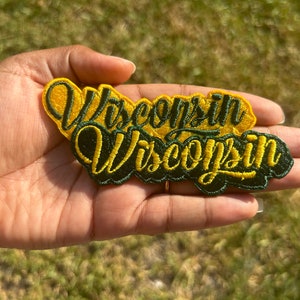 May include: A green and yellow embroidered patch with the word "Wisconsin" written twice in a stylized font.