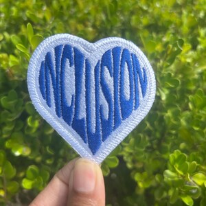 May include: A heart-shaped patch with the word "INCLUSION" embroidered in blue on a white background. The patch is surrounded by a white border and is set against a backdrop of green foliage.