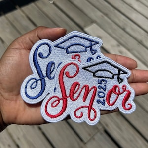 May include: Two embroidered patches with the word "Senior" in a cursive font, one in blue and the other in red. Each patch features a graduation cap and the year "2025". The patches are held in a hand against a wooden background.