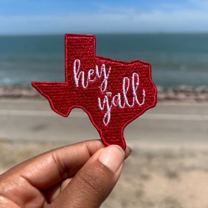 May include: A red embroidered patch in the shape of the state of Texas, with the words "hey y'all" in white script. The patch is held up against a blurred background of the ocean and sky.