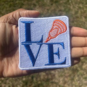 May include: Embroidered patch with the word "LOVE" in blue capital letters and an orange lacrosse stick on a light blue background. The patch is square with a white border.