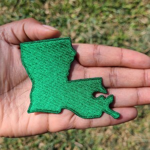 May include: A green embroidered patch in the shape of the state of Louisiana. The patch features a dense, textured stitch pattern throughout its surface, creating a solid green silhouette that mimics the geographic outline of the state.