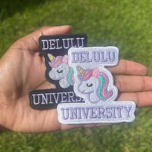 May include: Two embroidered patches featuring a unicorn head and the text DELULU UNIVERSITY. One patch has a black background with white text, while the other has a light purple background with dark purple text. Both unicorns have pink and teal manes.