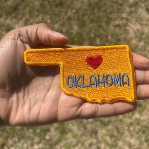 May include: Embroidered patch in the shape of Oklahoma, with a gold background and a red heart. The word "OKLAHOMA" is embroidered in blue. The patch is held in a hand.