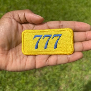 May include: A rectangular embroidered patch featuring a bright yellow background with a stitched border. The number 777 is centered on the patch in a bold, blue thread, creating a clear contrast against the yellow fabric surface.