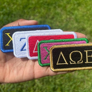May include: A set of five rectangular embroidered patches with Greek letters. The patches feature black, white, red, pink, and black backgrounds with contrasting borders and embroidered Greek characters including Delta, Omega, and Epsilon.