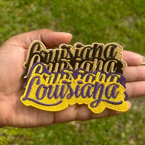 May include: Four embroidered patches with the word "Louisiana" in a cursive font. The patches are layered on top of each other, with the top patch being yellow with purple stitching, the second patch being black with yellow stitching, the third patch being yellow with black stitching, and the bottom patch being beige with black stitching.