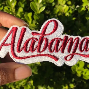 May include: White embroidered patch with the word "Alabama" in red cursive lettering.