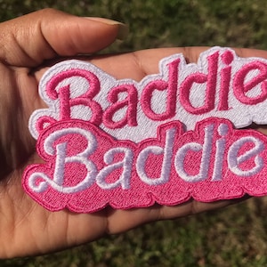 May include: Two pink and white embroidered patches with the word "Baddie" in white lettering. The patches are layered on top of each other.
