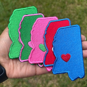 May include: A collection of embroidered patches in the shape of the state of Mississippi. The patches are in various colors: green, pink, red, and blue. Each patch features a heart, with the blue patch having a red heart.