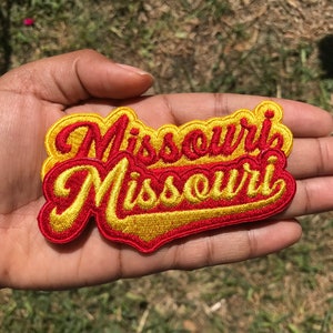 May include: A red and yellow embroidered patch with the word "Missouri" written twice in a stylised font.