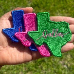 May include: Three embroidered iron-on patches shaped like the state of Texas. The patches are blue, pink, and green. The green patch has the word "Dallas" embroidered in pink thread.